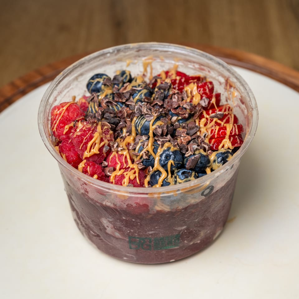 Berry smoothie bowl topped with raspberries, blueberries, cacao nibs, and peanut butter drizzle in a clear cup.