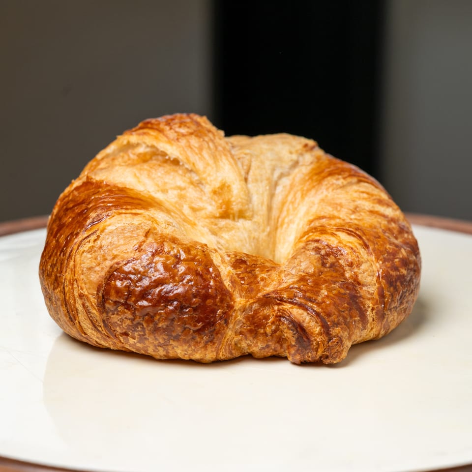 Golden-brown, flaky croissant-style pastry resting on a white plate against a dark background.